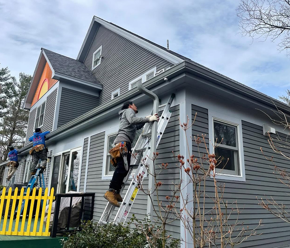 vermont-gutter-company-burlington-vt-1 Gray home in Burlington, VT, with a ladder propped against the side and a man repairing the gutters.