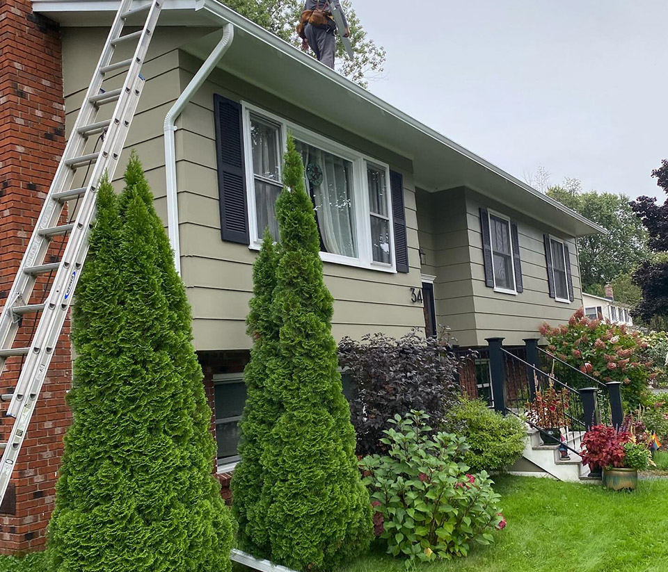 vermont-gutter-company-burlington-vt-3 Home in Burlington, VT, surrounded by green grass and shrubs, with a ladder along the side as people begin working on the gutters.