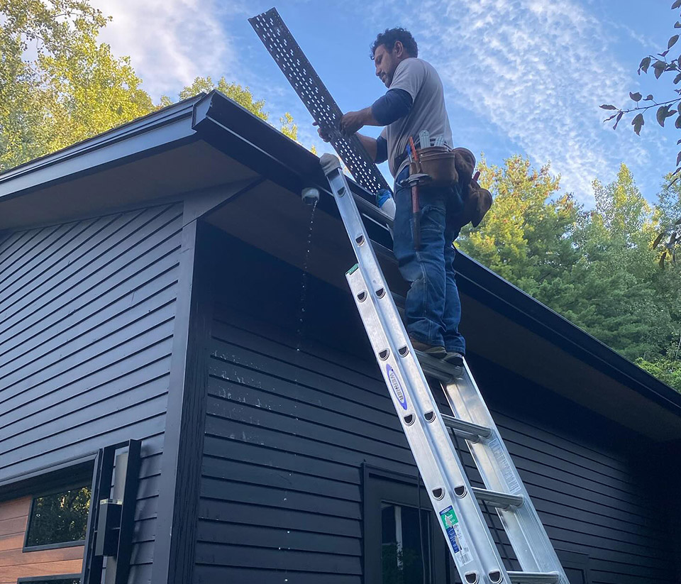 vermont-gutter-company-burlington-vt-5 Man standing on a ladder propped against a home in Burlington, VT, installing gutters on the roof.
