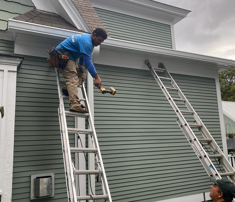 Two ladders propped against a home in Colchester, VT, with a man on one ladder preparing to work on the gutters.