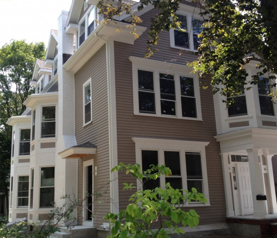 vermont-gutter-company-essex-junction-vt-3 Exterior of a tan-colored home in Essex Junction, VT with tall windows, neighboring homes nearby, and white gutters installed.