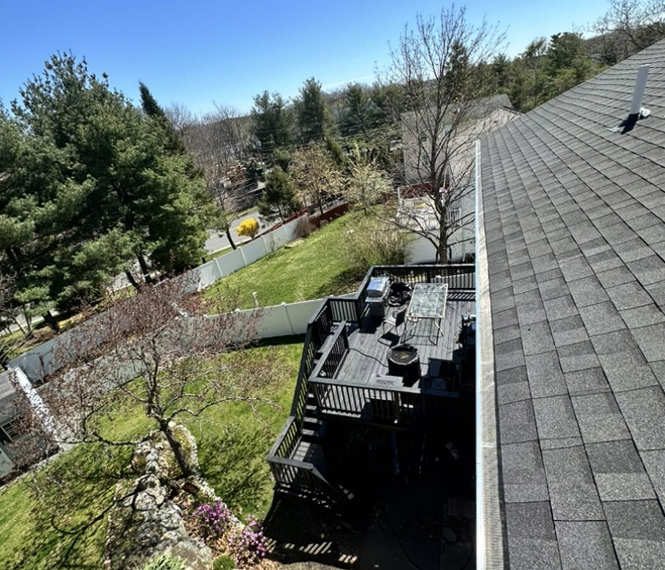 Top view of a shingled roof showing white gutters, a fenced yard with green grass, a tree, and a deck in Essex, VT.