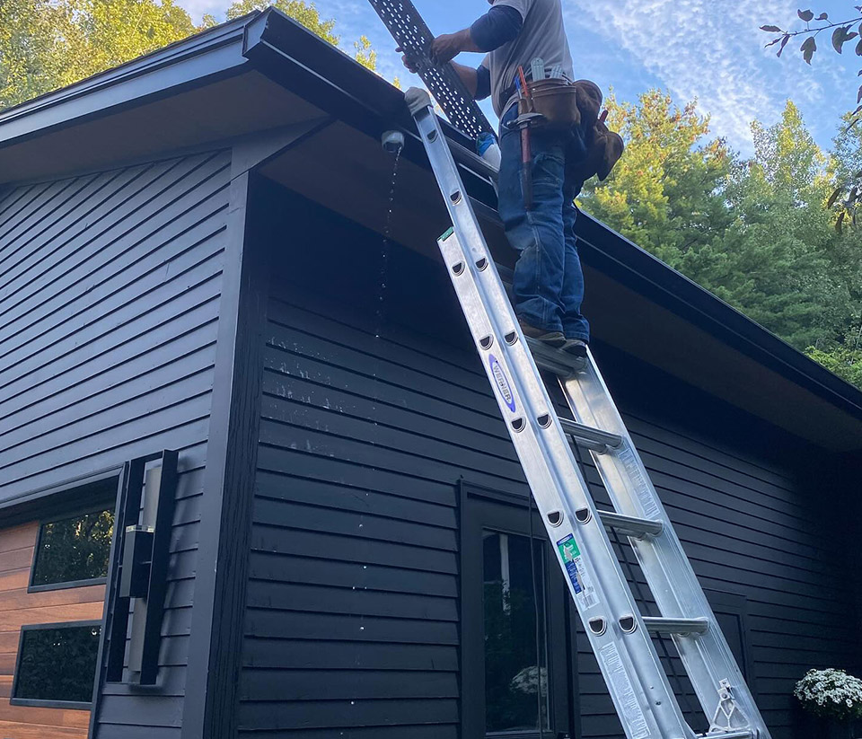 vermont-gutter-company-hartford-vt-4 Silver ladder propped against a garage with dark siding in Hartford, VT, with a person on the ladder working to install gutters on the home.