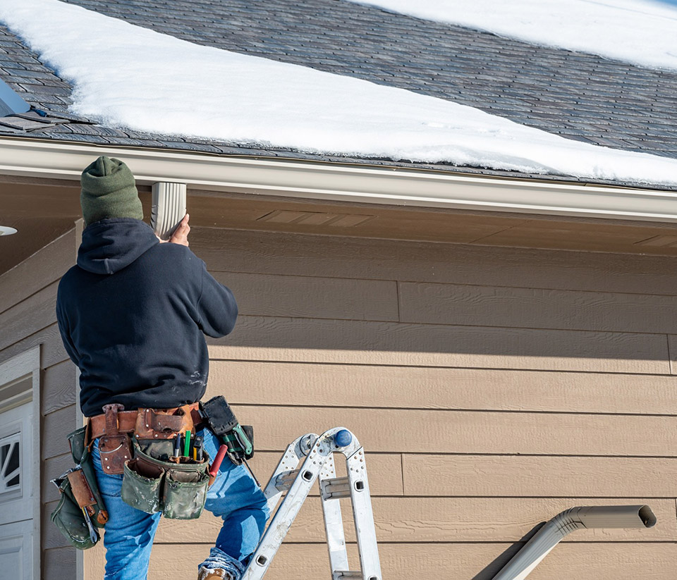 vermont-gutter-company-middlebury-vt-1 Man on a ladder against a tan home in Middlebury, VT, with snow on the roof, repairing the gutters.