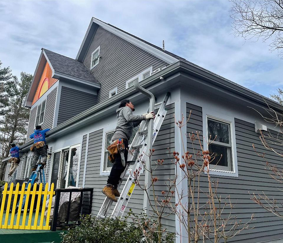 vermont-gutter-company-middlebury-vt-3 Man on a ladder against a grey home in Middlebury, VT, repairing the gutters.
