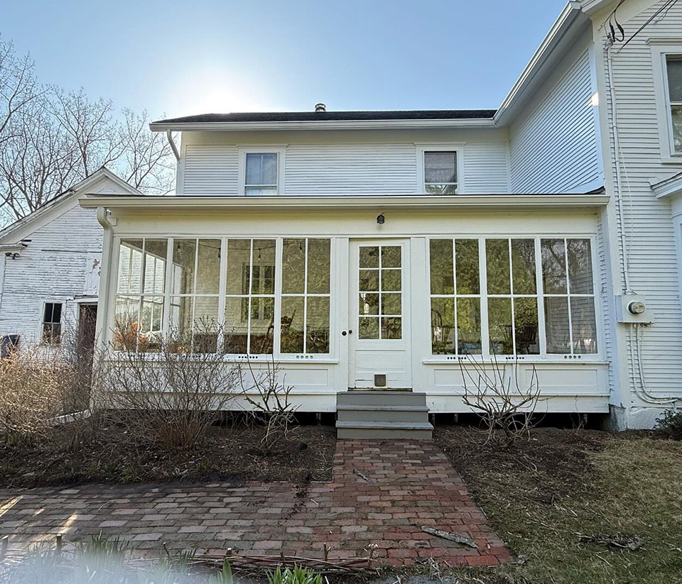 vermont-gutter-company-milton-vt-3 Exterior of a white home with an enclosed front porch and tall windows, a brick walkway leading to the entrance, and newly installed white gutters in Milton, VT.