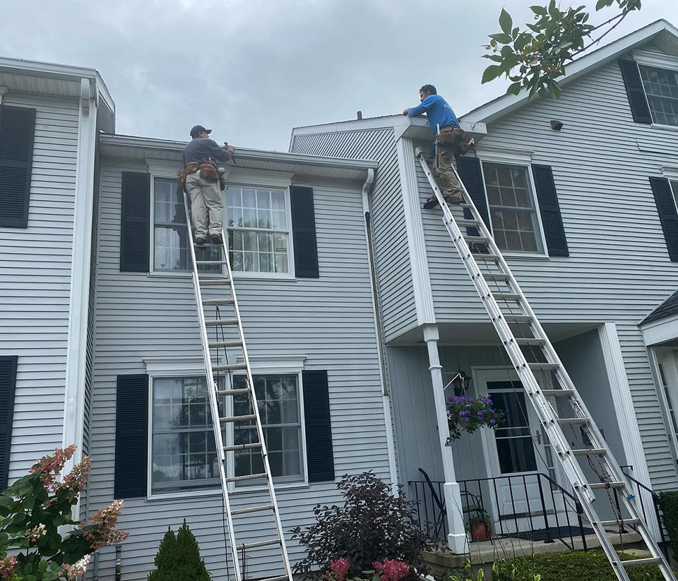 vermont-gutter-company-milton-vt-4 Exterior of a building in Milton, VT with two ladders set against the structure and two workers positioned on the ladders installing gutters along the roofline.