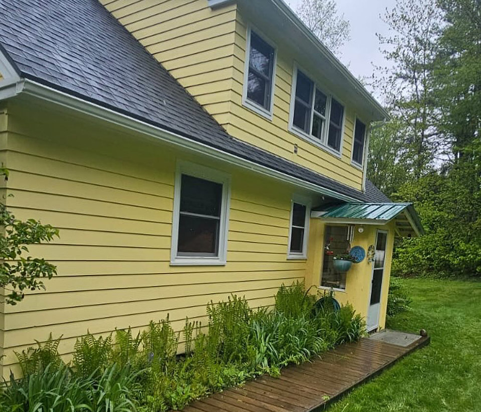 vermont-gutter-company-milton-vt-5 Side exterior of a yellow home in Milton, VT with a sidewalk leading to the door, surrounded by green grass and plants, and newly replaced gutters.