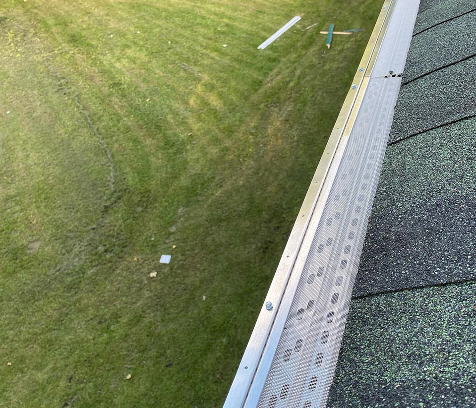 vermont-gutter-company-milton-vt-6 Close-up of a gutter with a leaf guard installed along a shingle roof, with green grass in the background in Milton, VT.
