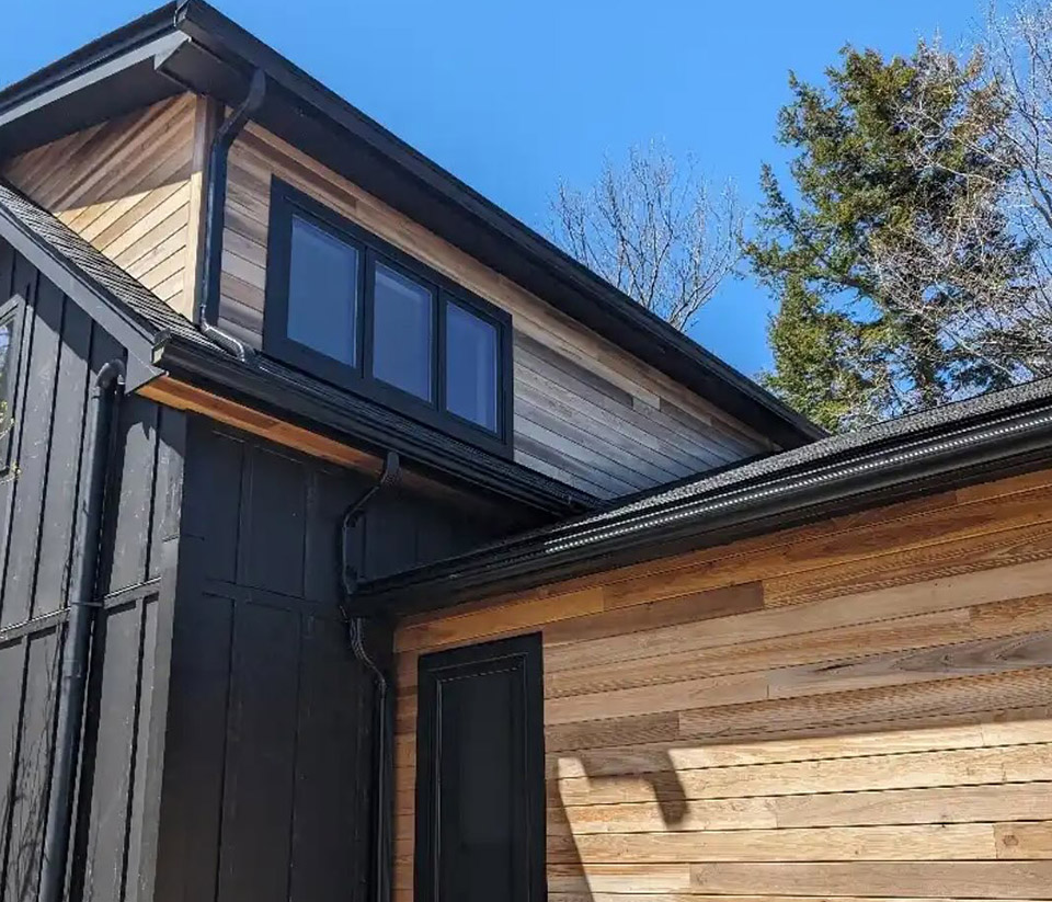 vermont-gutter-company-rutland-vt-1 Close‑up of the side of a home in Rutland, VT, with dark wood and natural wood siding, showing dark‑colored gutters.
