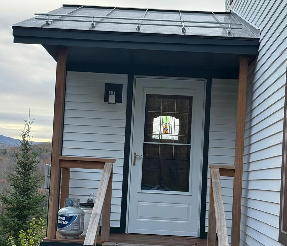 vermont-gutter-company-shelburne-vt-4 Front porch close‑up of a home in Shelburne, VT, showing the wooden deck and metal roof with trees in the background.