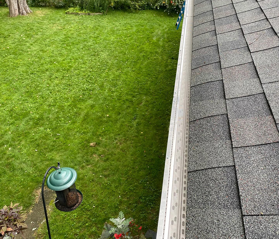 vermont-gutter-company-shelburne-vt-6 Close-up of white gutters on the side of a home in Shelburne, VT, showing the shingle roof, surrounding grass, and a bird feeder.