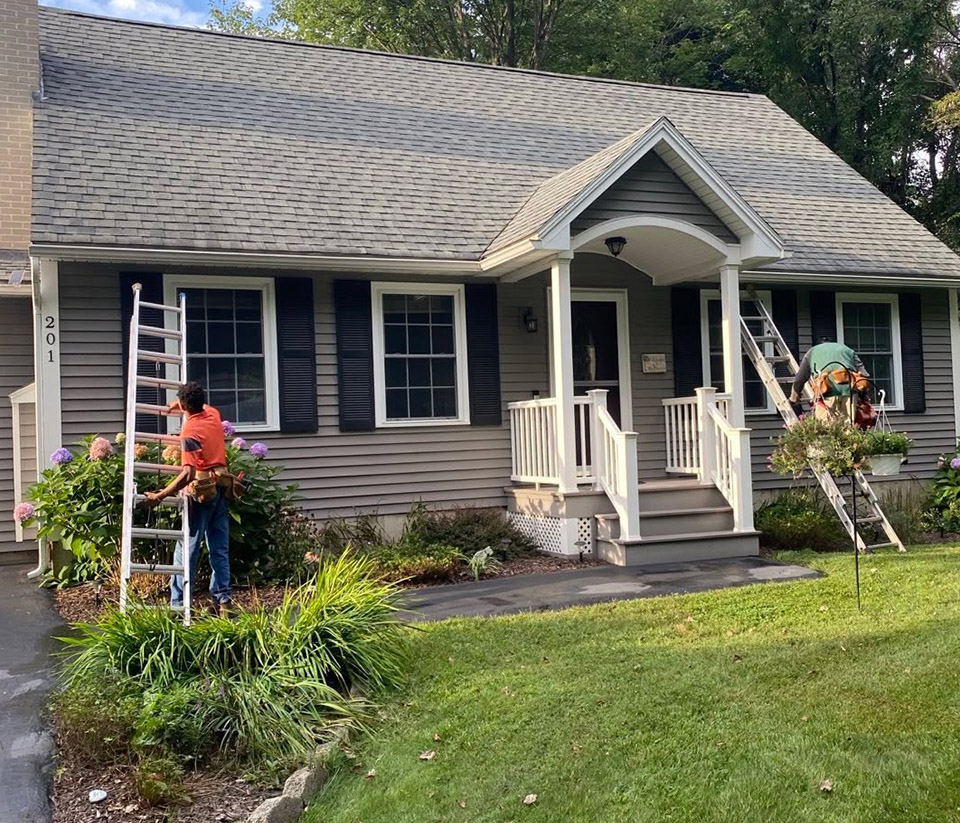vermont-gutter-company-south-burlington-vt-4 Grey home in South Burlington, VT, with two men propping ladders against it, preparing to work on the gutters.