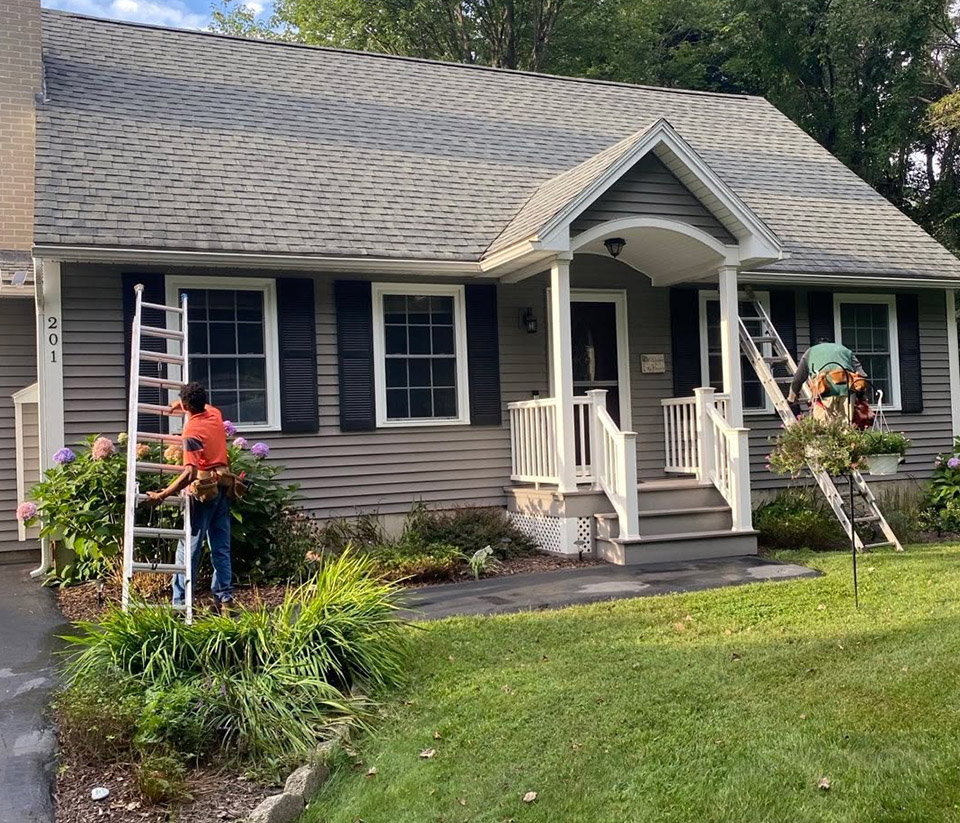 vermont-gutter-company-st-albans-vt-5 A man in St. Albans, VT, with a ladder against a grey home with a small white porch and green grass, preparing to work on the gutters.