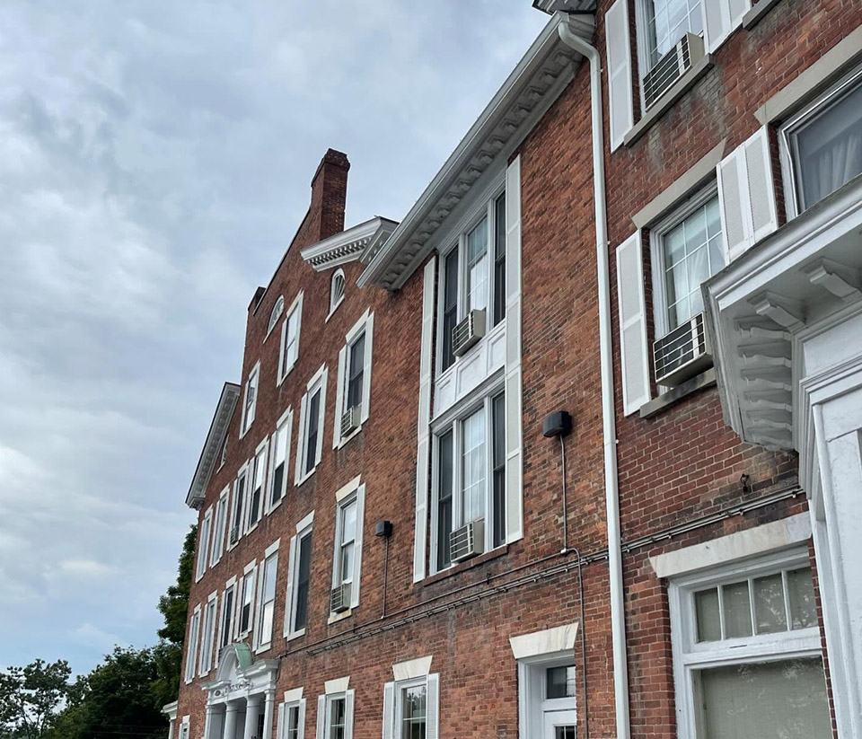 vermont-gutter-company-st-johnsbury-vt-1 Red brick multi-story building with white trim, tall windows, and several window air conditioning units under a cloudy sky in St. Johnsbury, VT.
