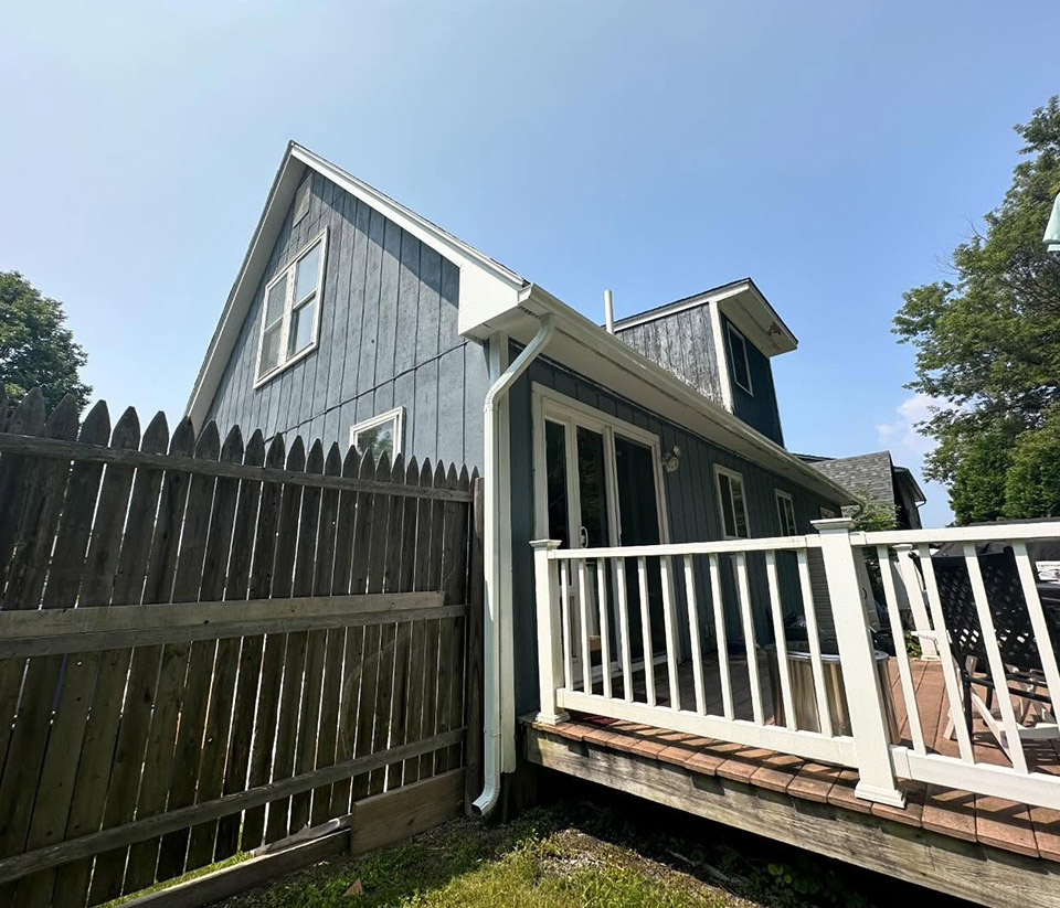 vermont-gutter-company-stowe-vt-1 Side view of a blue home in Stowe, VT showing white gutters along the roofline, a wooden fence, and a deck with white railing.