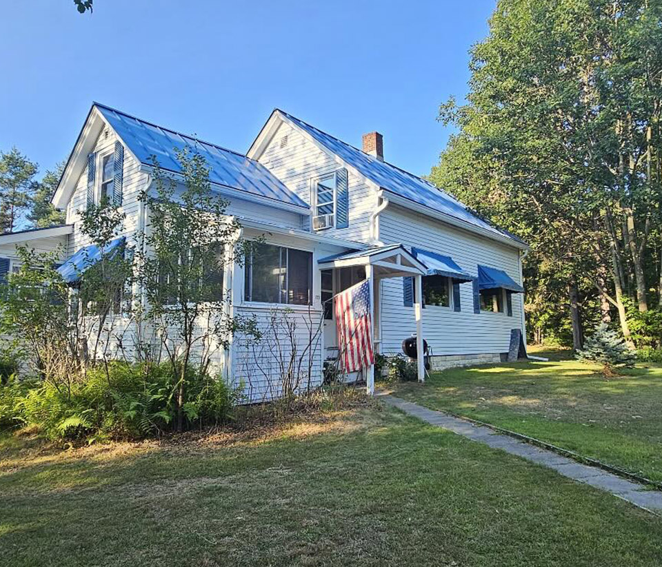 vermont-gutter-company-stowe-vt-3 White home in Stowe, VT, with an American flag attached, surrounded by green grass and trees, with newly installed white gutters.