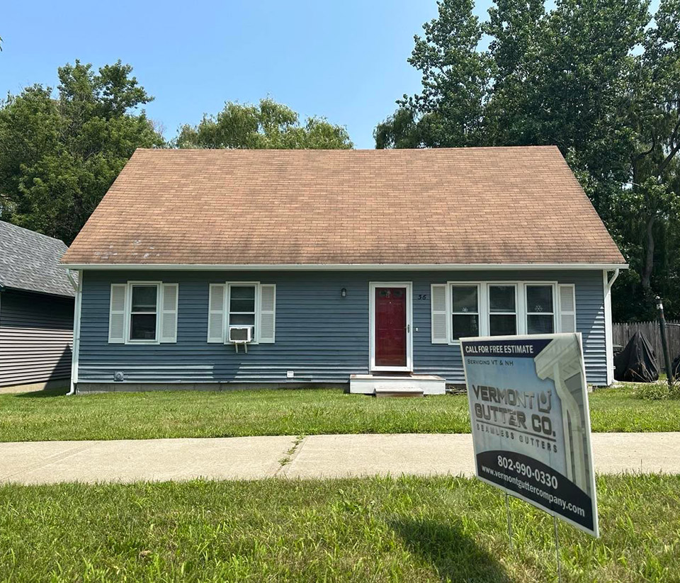 vermont-gutter-company-stowe-vt-5 Front of a blue home in Stowe, VT with a slanted roof, green grass, and a sign in the yard.