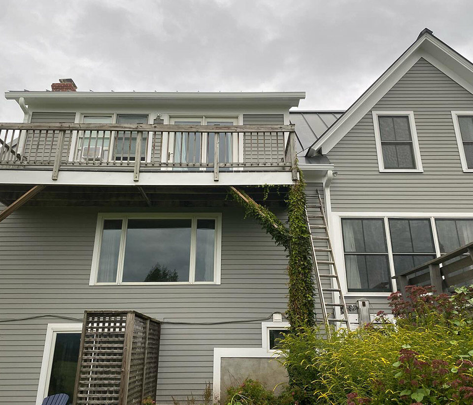 vermont-gutter-company-swanton-vt-4 Home in Swanton, VT, with light green siding, a wooden deck, white window trim, and a ladder propped against it as someone prepares to work on the gutters.