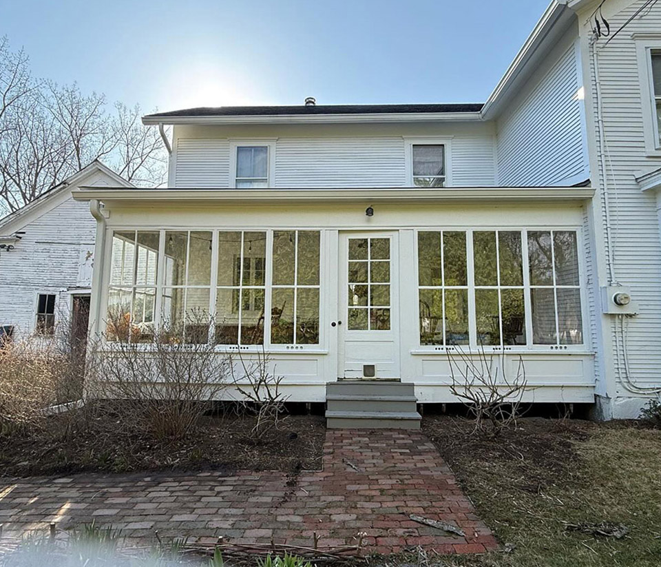 vermont-gutter-company-waterbury-vt-1 Front of a white home in Waterbury, VT, with an enclosed porch, windows, a stone brick walkway leading to the door, and white gutters.