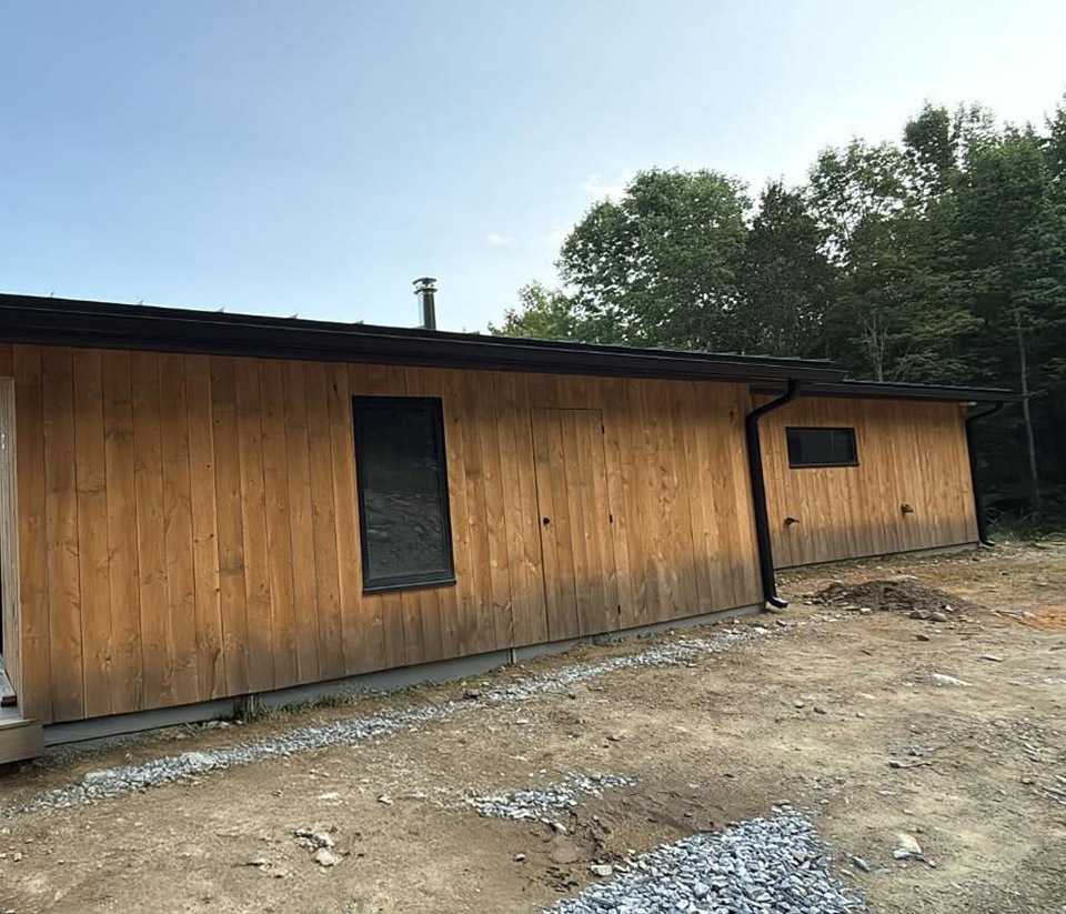 vermont-gutter-company-williston-vt-3 Exterior of a home in Williston, VT, featuring natural wood siding, two windows with black trim, newly installed black gutters, and surrounding green trees.
